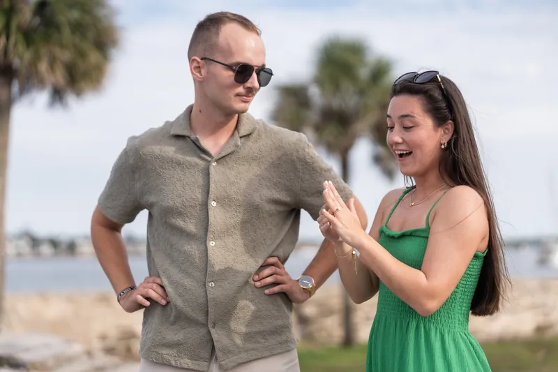 Two people stand together on a beach with palm trees; the woman in green gestures expressively while the man watches.