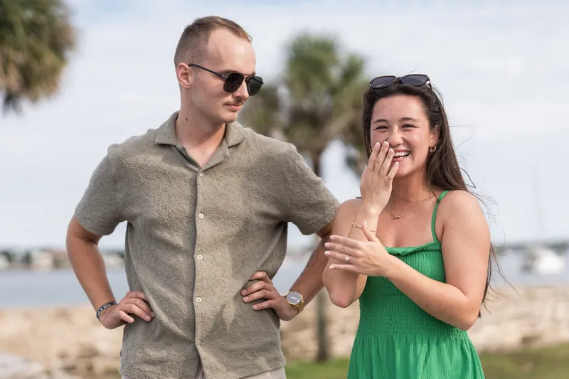 Woman in a green dress grins while touching the man's chest as he stands with hands on hips, both near the waterfront.