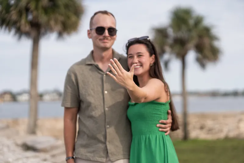 Couple poses together on the beach with the woman holding up her hand to display a ring, palm trees and water behind them.