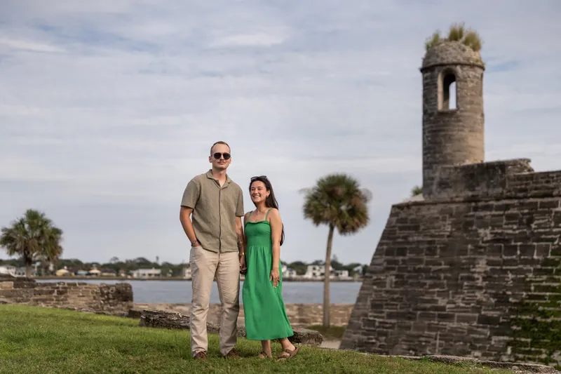 Two people stand on a grassy area in front of a historic stone tower with water and palm trees in the background.