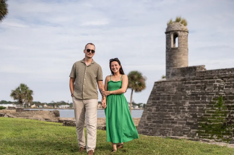Two people walk hand-in-hand across a grassy field with a historic stone tower and fortifications in the background.
