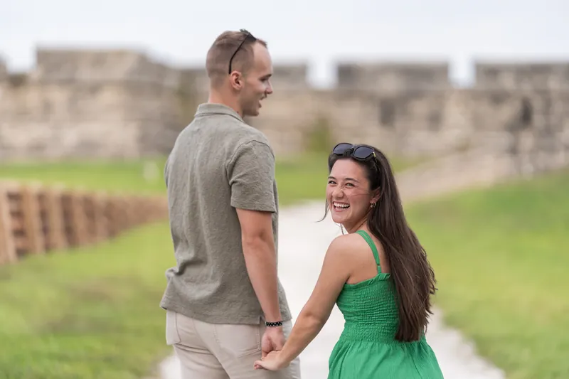 A couple holds hands while walking on a path, both smiling as they look at each other with stone ruins behind them.