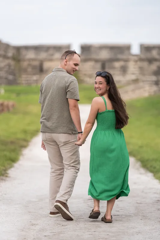 Man and woman walk together down a tree-lined path, holding hands and looking at each other with old fortification walls visible ahead.