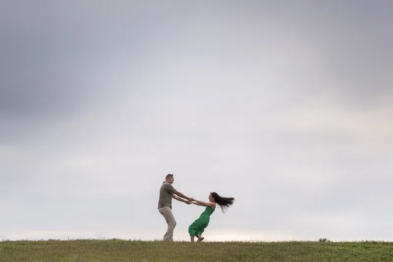 Two people spin playfully across an open grassy field under an overcast sky, one in a khaki shirt and the other in a green dress.