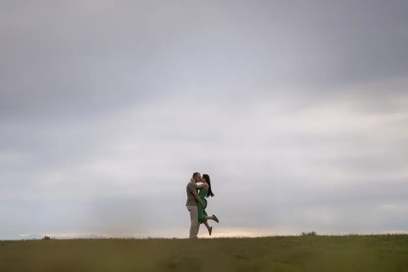 Someone carries their partner across a wide open field, both framed against a cloudy horizon and empty landscape.