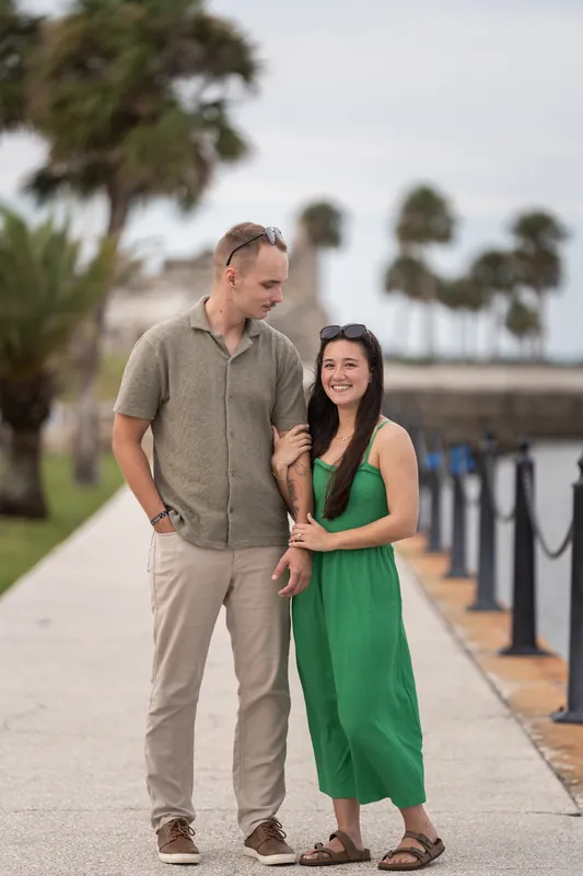 A man in khaki and a woman in green stand close together on a waterfront promenade with palm trees and historic structures along the shore.