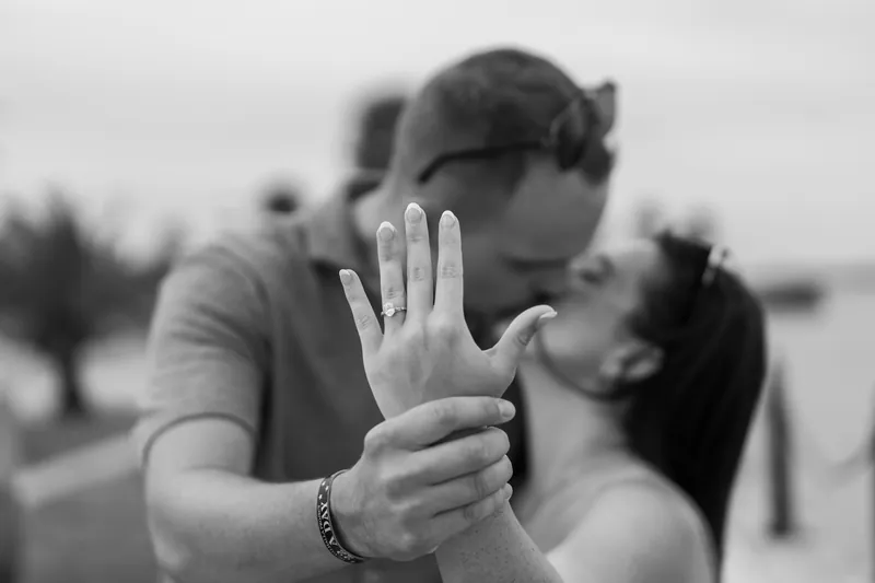 Two people kiss in black and white while one holds up their hand displaying a ring.