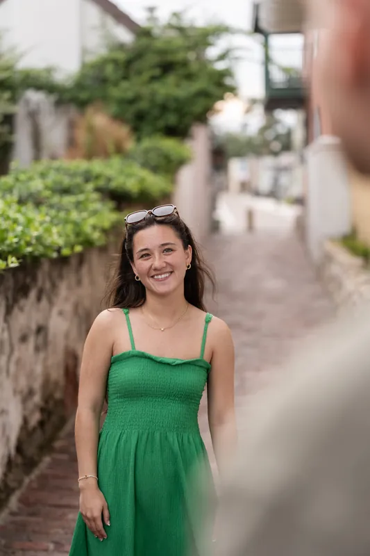Person in a green dress smiles at the camera on a brick pathway with greenery behind.