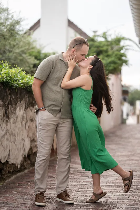 Two people kiss on a paved walkway beneath architectural elements and climbing vines.