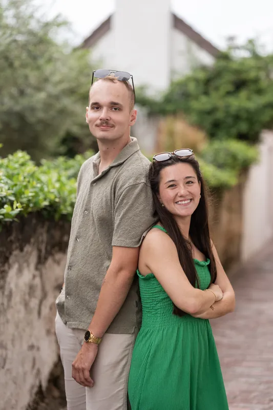 Couple stands back-to-back on a brick path, both wearing sunglasses on their heads and smiling.
