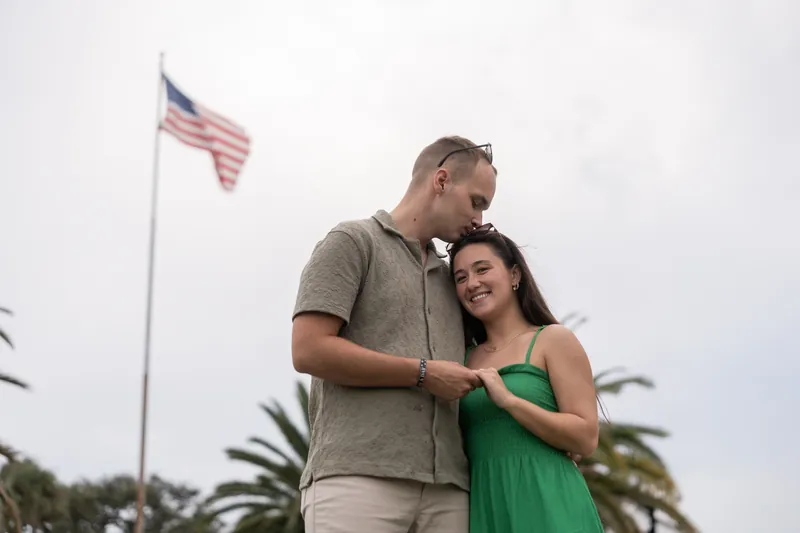 Couple stands together under an American flag against an overcast sky with palm trees nearby.