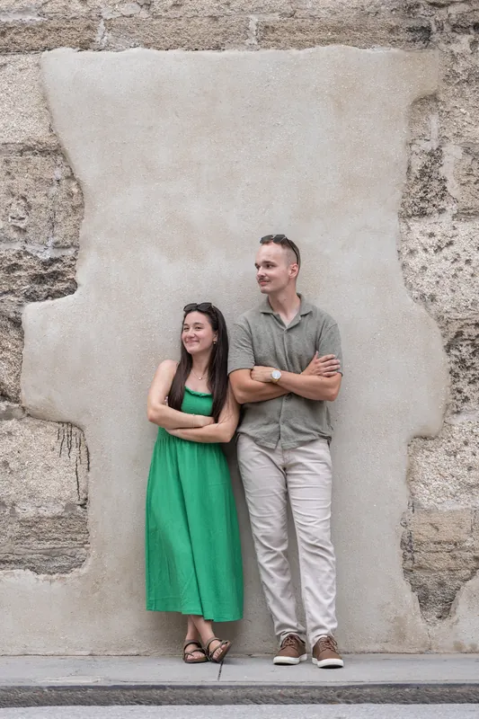 Mason and Caitlin stand side by side against a textured stone wall, both in casual attire with arms crossed.
