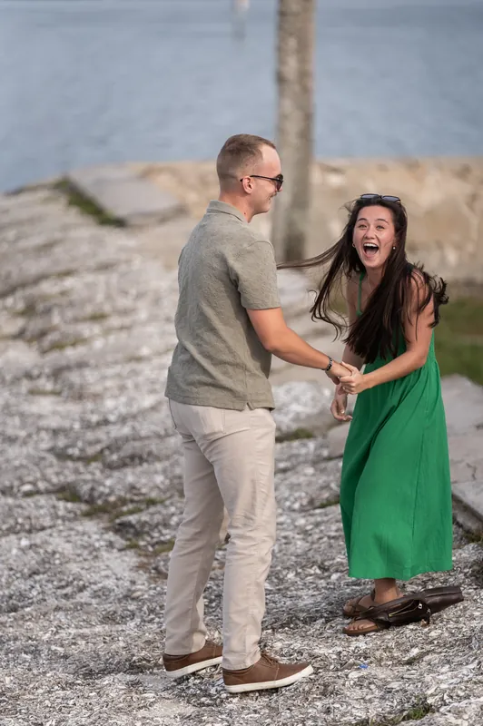 Two people stand on a pebbly shore, one in a gray shirt turning toward the other in a green dress who laughs.