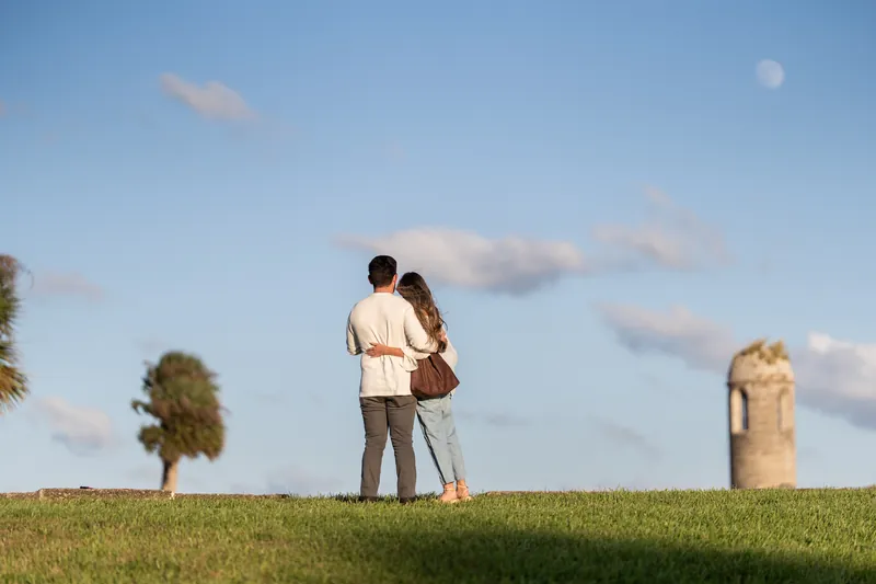 Two people stand together on a grass field at Castillo de San Marcos with historic stone fortification and palm trees under a blue sky.