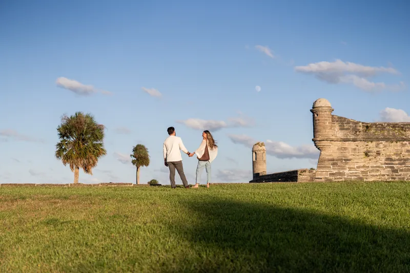 A couple walks across the lawn at Castillo de San Marcos toward the stone fortress walls, with palmetto trees dotting the grassy grounds.