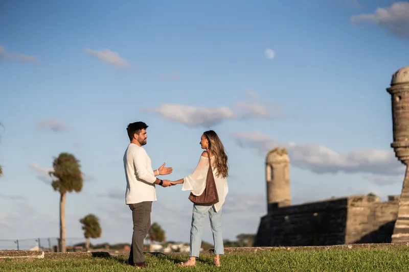 A person in a light shirt kneels on one knee facing their partner on the grass, with the historic stone fort and palm trees in the background.