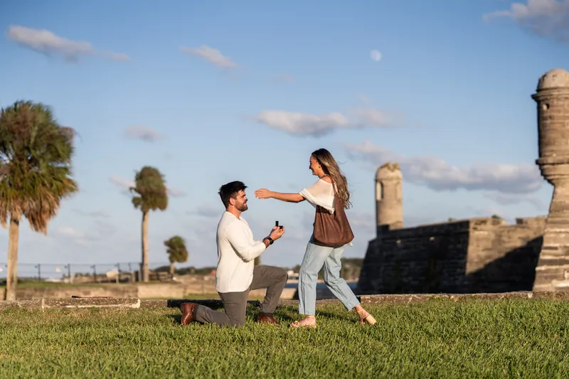 A person on one knee presents a ring to their partner during a proposal moment on the grounds of Castillo de San Marcos.