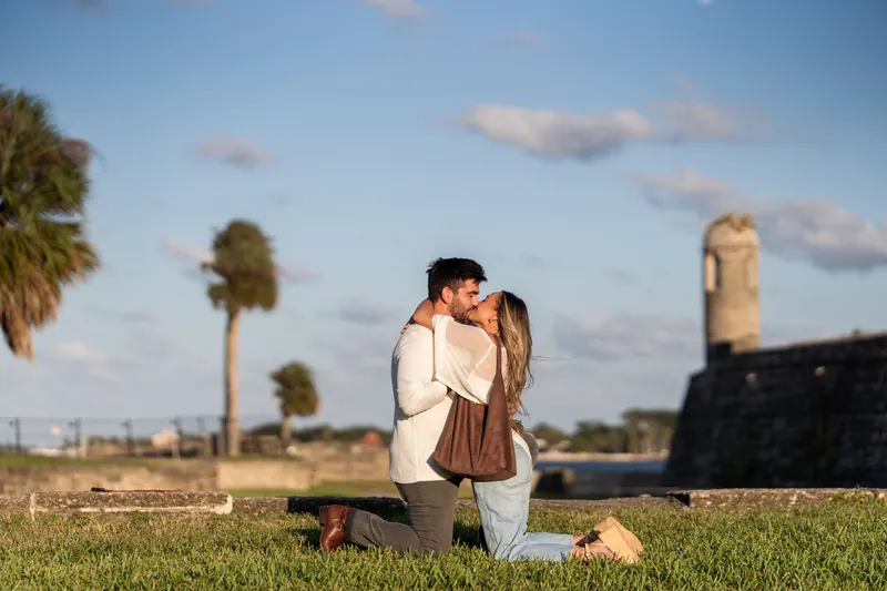 Matt and Mary embrace on the grass at Castillo de San Marcos with palm trees and the fort's stone walls behind them under a blue sky.
