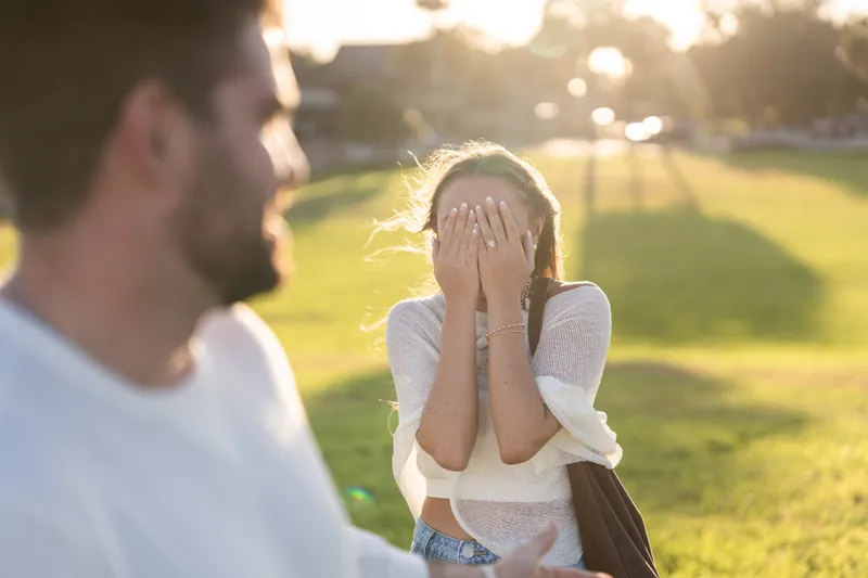 A person covers their face with both hands in shock while their partner looks on at the historic fort's grassy grounds.