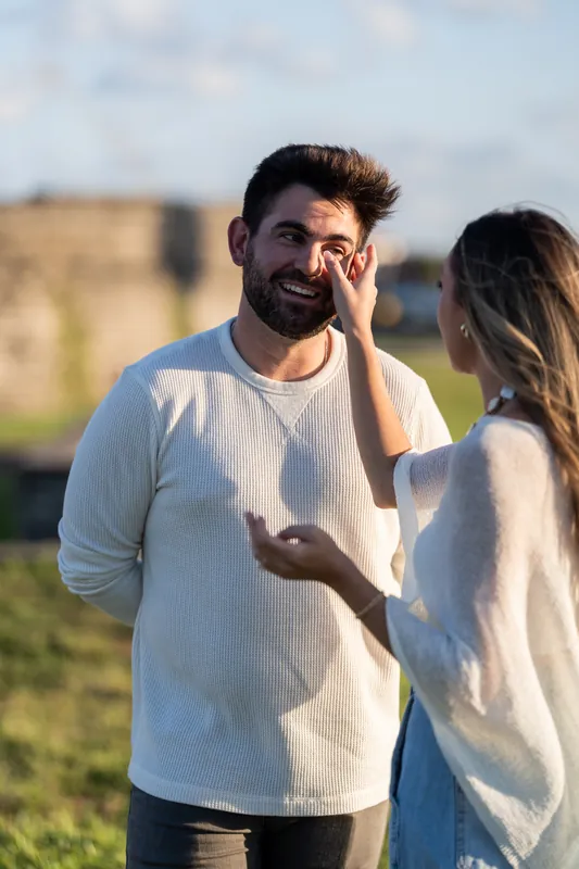 A man in a cream-colored shirt touches his face pensively while his partner stands beside him on the fort's lawn.