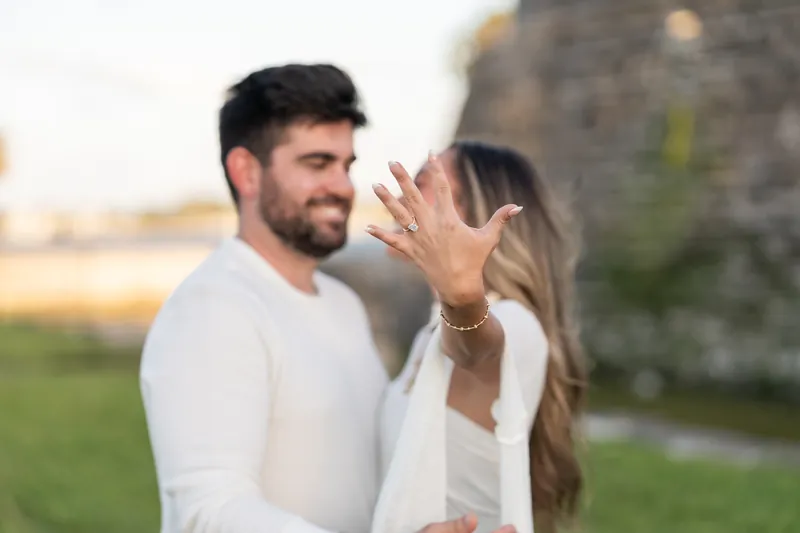 A person in a white long-sleeve shirt displays their engagement ring while standing in front of historic fort architecture.