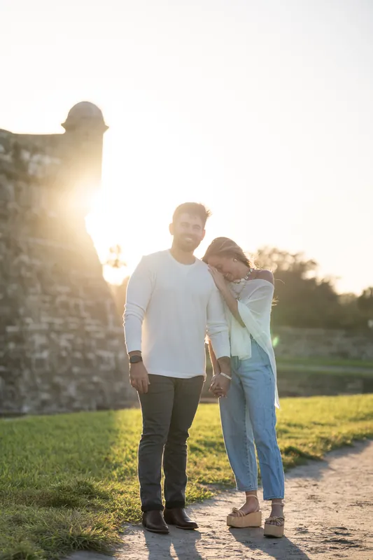 Two people walk hand-in-hand along a path beside the Castillo de San Marcos as sunlight filters through the stone structure.