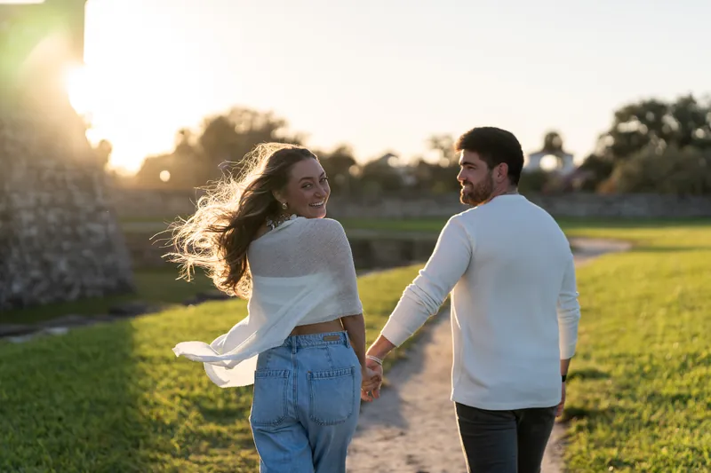 Two people in casual wear walk along a grassy path with backlit sunset and the fort's stone wall framing the left edge.
