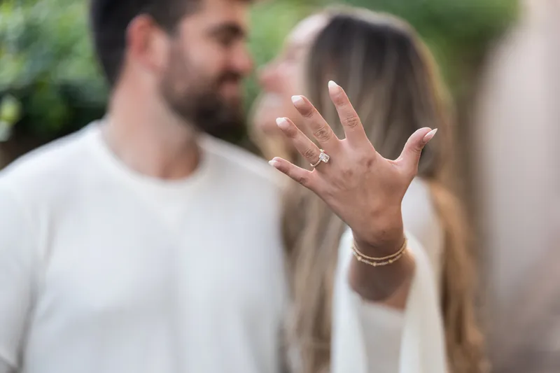 A hand displays a diamond engagement ring, with Matt visible slightly out of focus in the background on a downtown St. Augustine street.