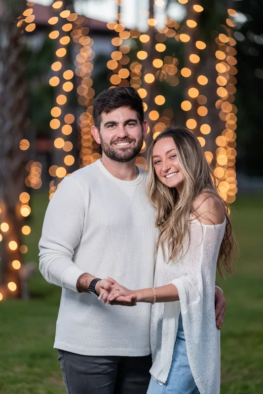 Matt and Mary smile at the camera with warm string lights glowing softly behind them in an evening garden setting.