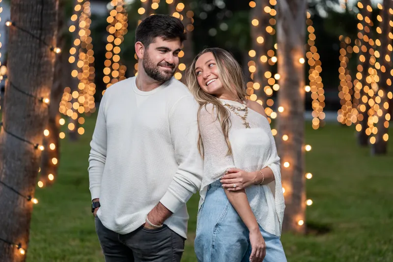 The couple stands together beneath draped golden lights strung between trees, both wearing cream and light blue layers.