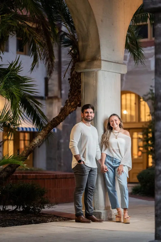 Two people hold hands walking through a palm-lined arched courtyard with warm-lit doorways at the Lightner Museum.