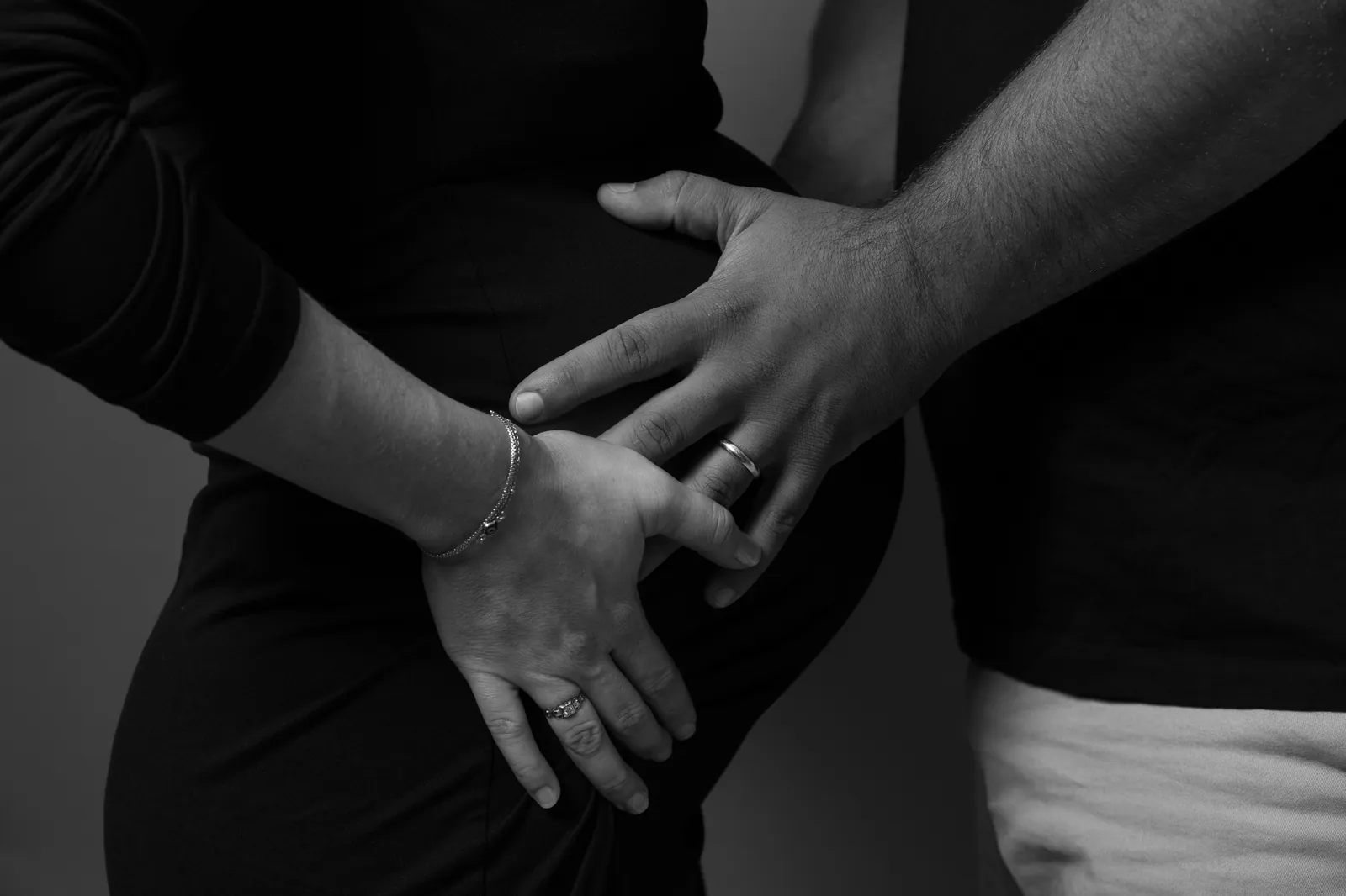 Detail shot of couple's hands with wedding rings resting on baby bump, black and white.