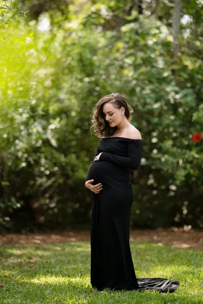 Expectant mother in garden setting, looking down at baby bump during outdoor maternity portrait.