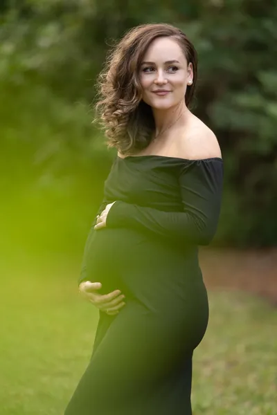 Maternity portrait with beautiful green bokeh background, expectant mother smiling outdoors.