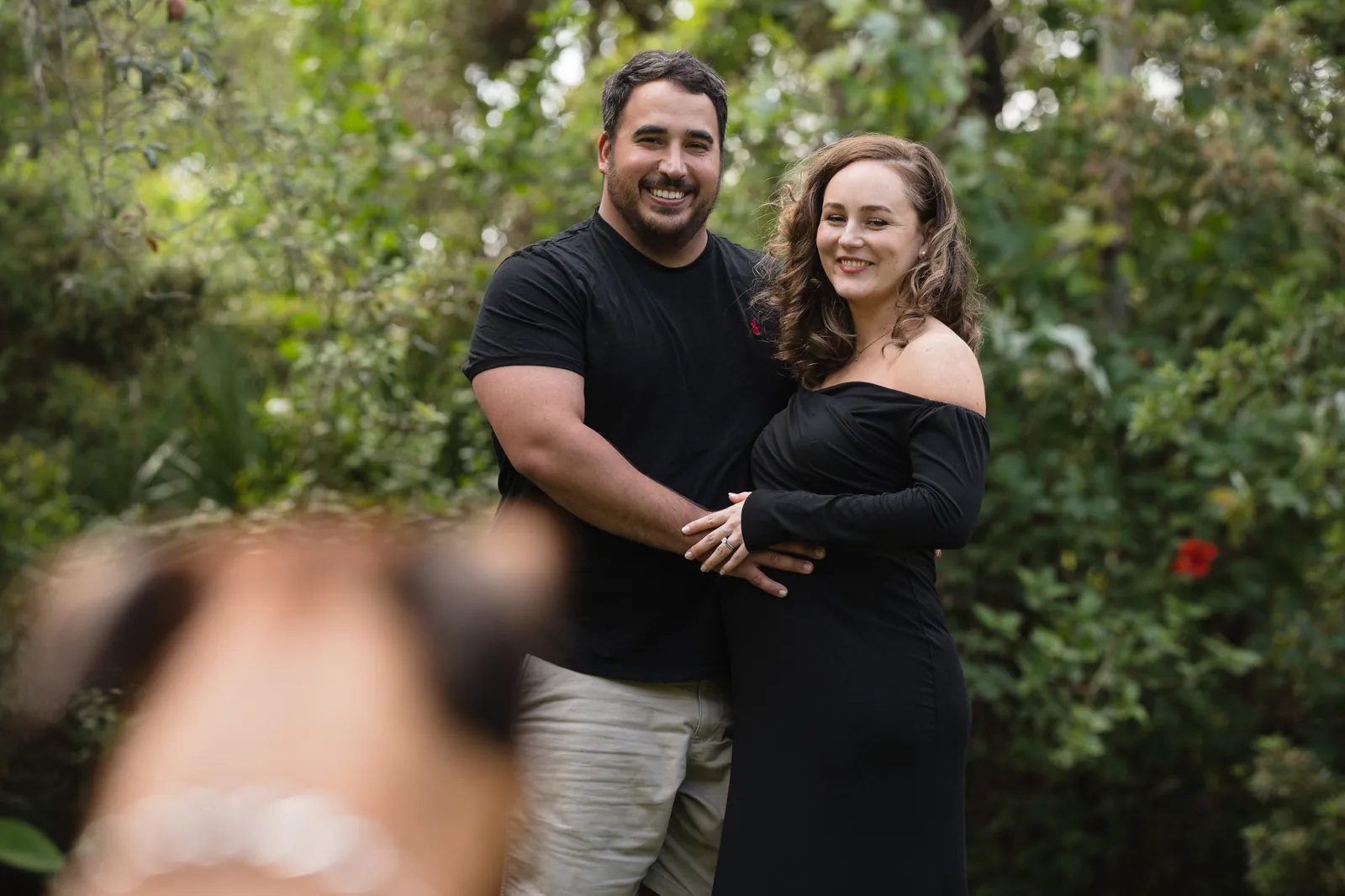Boxer dog photobombing couple's maternity portrait in the garden, everyone smiling.