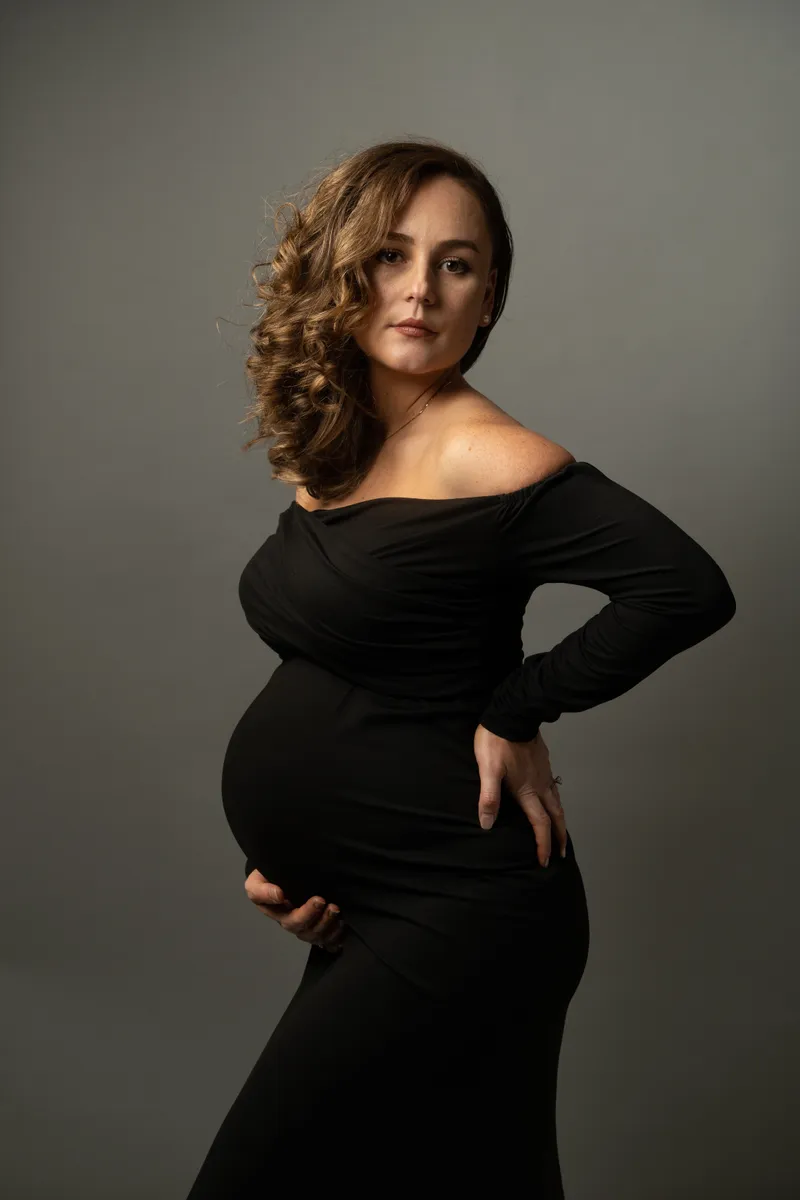 Megan in a black long-sleeve maternity dress, hand supporting her belly, gazing thoughtfully to the side.