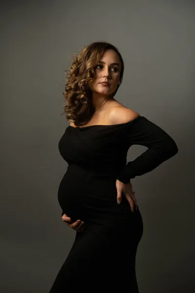 Elegant studio maternity portrait, expectant mother in black dress looking away from camera.