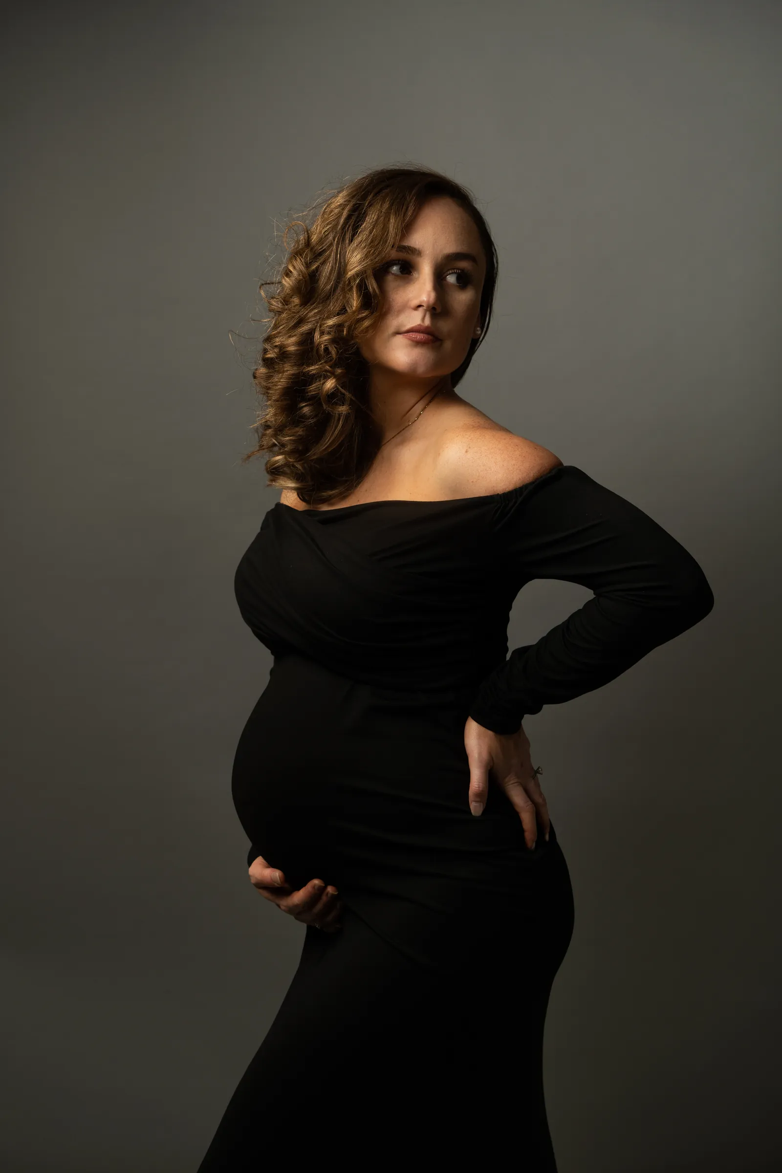 Elegant studio maternity portrait, expectant mother in black dress looking away from camera.