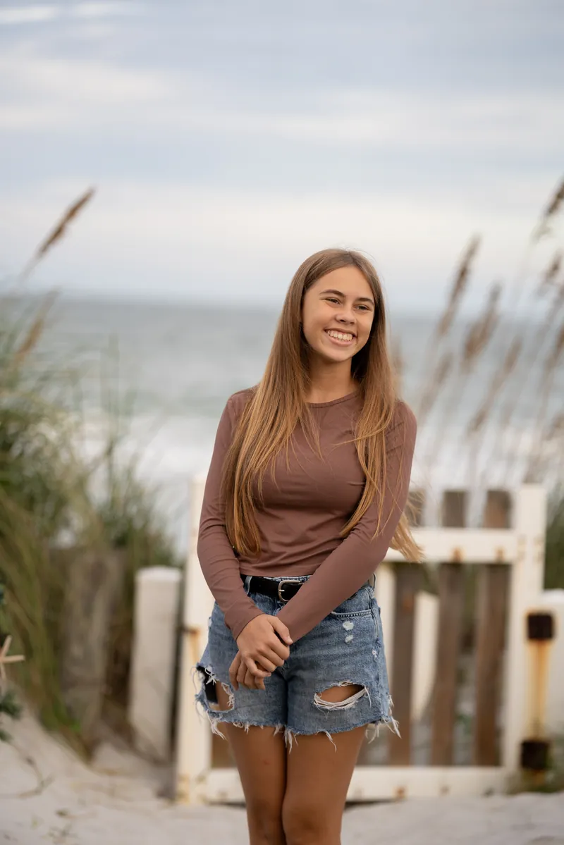 Megan smiles at Vilano Beach wearing a brown long-sleeve top and distressed denim shorts, ocean and dune grasses behind her.
