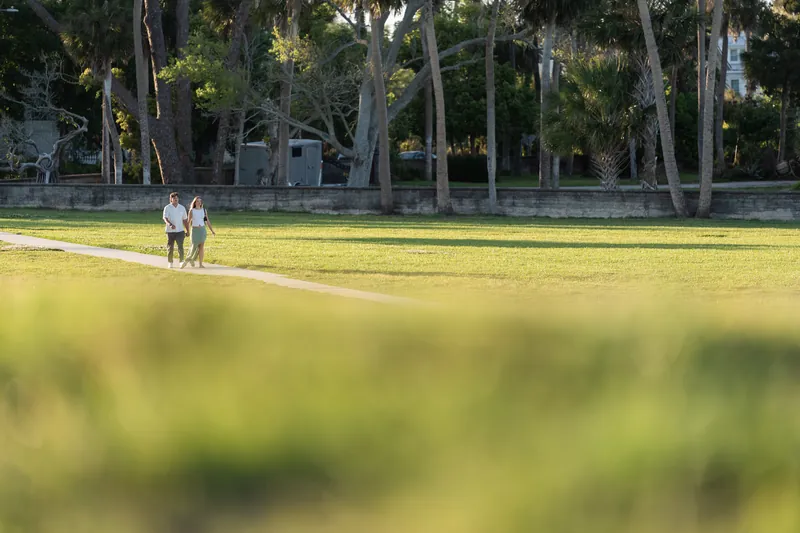 Michael and Katie hold hands while walking across a sunlit grass field lined with tall trees.