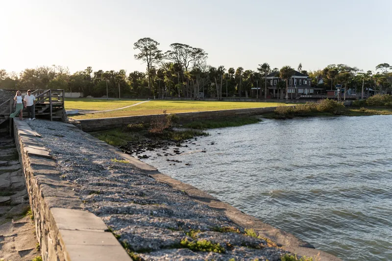 A waterfront park with a wooden seawall, manicured lawn, and tree-lined shore under afternoon light.