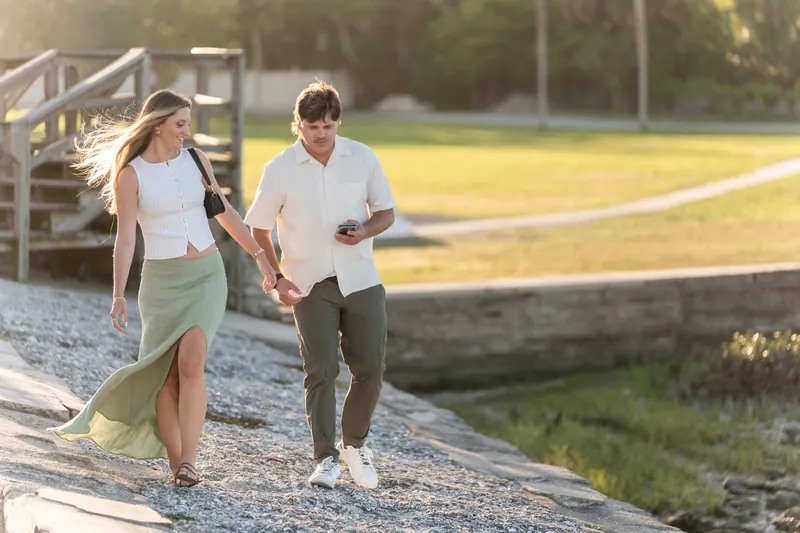 Katie in a sage green skirt and Michael in a white shirt walk hand-in-hand on a gravel path by the water.