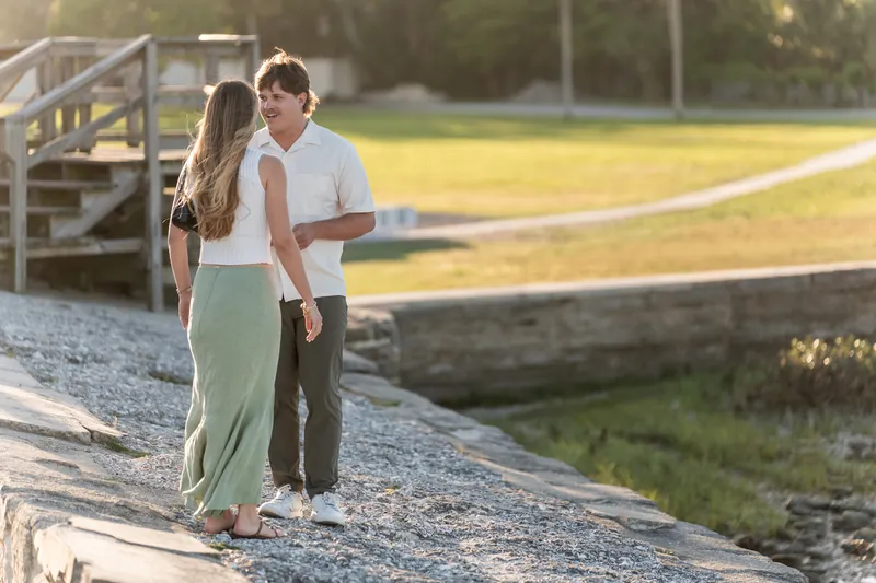 Michael kneels on one knee to propose to Katie on a waterfront path, with wooden dock and grass field behind.
