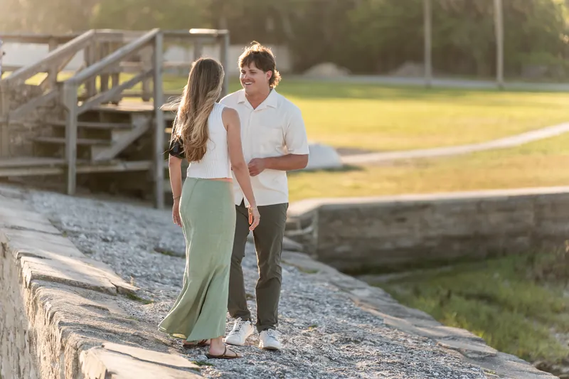 Katie holds her hand up to her chest in surprise as Michael proposes on the gravel path beside the water.