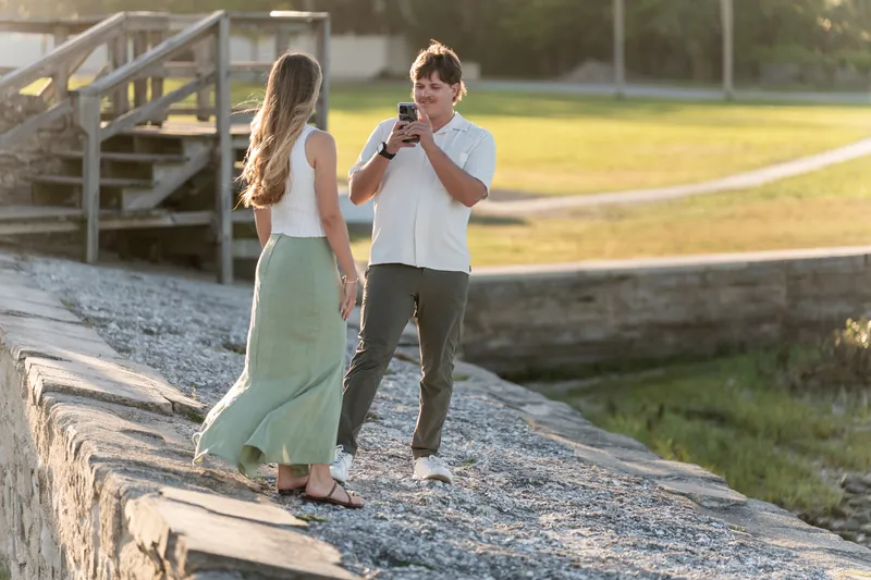 Katie and Michael embrace after his proposal on the waterfront path, with the dock and lawn in the background.