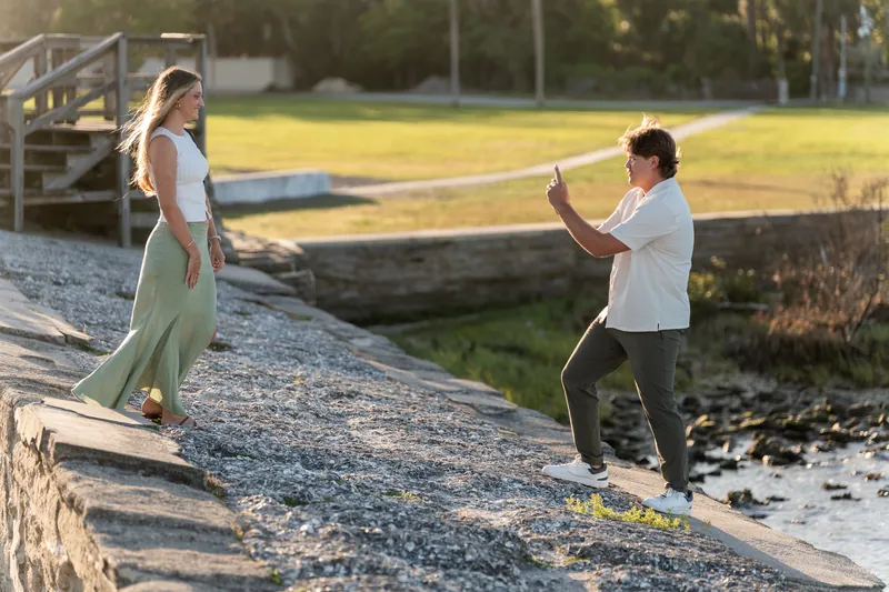 Michael kneels on gravel while Katie stands nearby as he proposes by the water at sunset.