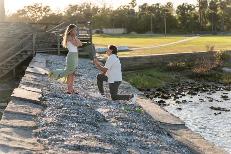 Katie laughs with her hand to her chest while Michael holds the ring box during their proposal on the rocky shore.