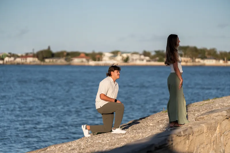 Michael kneels on a boulder overlooking blue water as he proposes to Katie in her sage green dress.