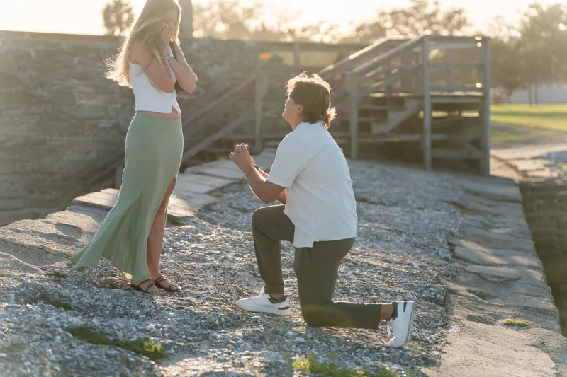 Katie's hand touches her chest in surprise as Michael proposes on his knee at the waterfront.