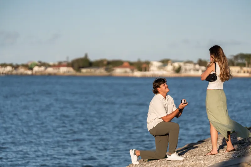 Michael kneels with the ring while Katie stands before him on the rocky shore at sunset.